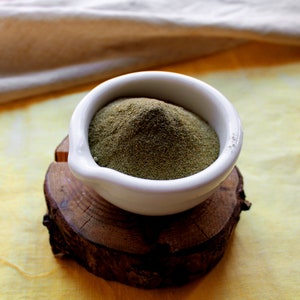 Può includere: Una ciotola di ceramica bianca piena di polvere verde è appoggiata su un piccolo supporto di legno rustico. La ciotola è centrata nell'inquadratura, con la polvere che trabocca leggermente dal bordo. Lo sfondo è un tessuto giallo tenue.