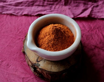 May include: A white ceramic mortar and pestle filled with a mound of bright orange powder, sitting on a wooden base against a pink fabric background.