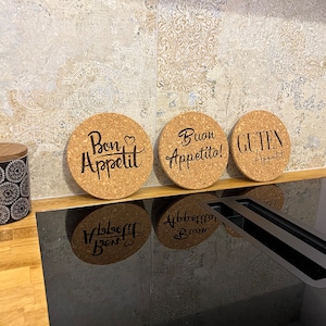 Könnte beinhalten: Drei runde Untersetzer aus Kork mit den Schriftzügen "Bon Appetit", "Buon Appetito!" und "Guten Appetit" in schwarzer Schrift. Die Untersetzer sind auf einer Holzoberfläche angeordnet, mit einem Hintergrund aus gemusterten Fliesen.
