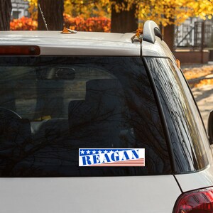 May include: A white car with a blue, red, and white sticker on the back window that says "REAGAN" with stars above the text.