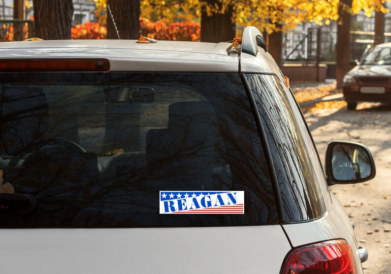 May include: A white car with a blue, red, and white sticker on the back window that says "REAGAN" with stars above the text.