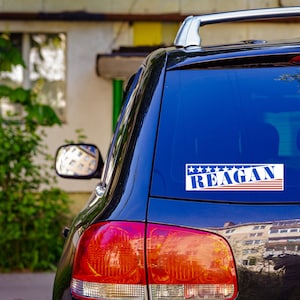 May include: A black car with a blue and red sticker on the back window that says "Reagan" with stars above the text.