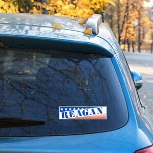 May include: A blue car with a sticker on the back window that says "REAGAN" in blue letters with red and white stripes. The sticker has five stars above the text.