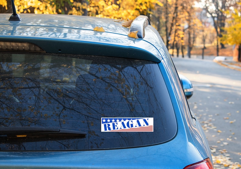 May include: A blue car with a sticker on the back window that says "REAGAN" in blue letters with red and white stripes. The sticker has five stars above the text.