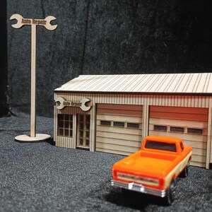 Könnte beinhalten: Miniatur-Modell einer Holz-Autowerkstatt mit einem orangefarbenen Pickup-Truck. Die Werkstatt hat zwei Garagentore und ein Schild mit der Aufschrift "Auto Repair". Ein hohes Schild mit einem Schraubenschlüssel-Design steht daneben.