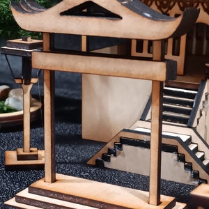 May include: A wooden model of a traditional Japanese gate, or torii, with a curved roof and two pillars. The gate is made of light brown wood and has a dark brown base.