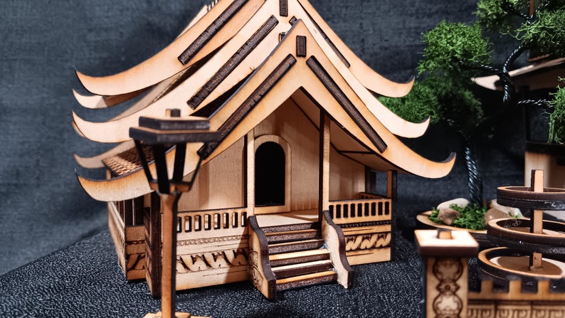 May include: A wooden model of a traditional Asian house with a curved roof and intricate details. The house has a small porch and a lamppost in front. The model is made of light brown wood and has a natural finish.