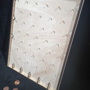 Könnte beinhalten: Ein hölzernes Plinko-Spielbrett mit einem dunkelbraunen Rahmen. Die hellfarbene Holzoberfläche hat zahlreiche kleine Stifte, die in Reihen angeordnet sind. Mehrere Cent-Münzen sind in der Nähe des unteren Randes des Bretts verstreut, was auf ein Spiel hindeutet. Das Brett ist abgewinkelt und zeigt seine Tiefe.