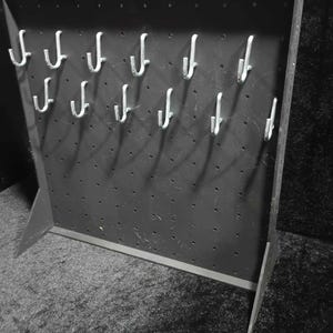 Op de afbeelding: Een zwarte displaystandaard met meerdere zilveren haken in rijen gerangschikt. De standaard heeft een reeks kleine gaatjes en is ontworpen om items vast te houden. De standaard is gemaakt van een donker materiaal en heeft een eenvoudig, functioneel ontwerp.