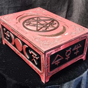 May include: A rectangular, red wooden box with intricate carvings. The lid features a circular design with a star-like pattern, while the sides display crescent moons and alchemical symbols. The box has a black interior and is likely used for storage.