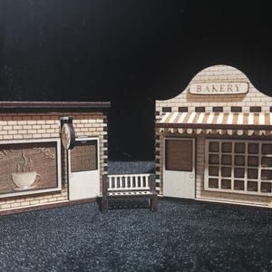 May include: Miniature wooden coffee shop and bakery models. The coffee shop has a sign that says "Coffee Shop" and a coffee cup graphic. The bakery has a striped awning and the word "Bakery" on top. A small bench sits between the two buildings.