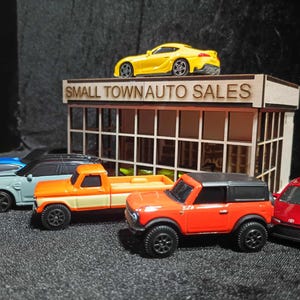 Könnte beinhalten: Eine Sammlung von Spielzeugautos, darunter ein gelber Sportwagen, ein orangefarbener Pickup und ein roter SUV, werden vor einem Miniatur-Holzgebäude mit dem Text "SMALL TOWN AUTO SALES" ausgestellt. Die Autos sind auf einer dunklen Oberfläche angeordnet.