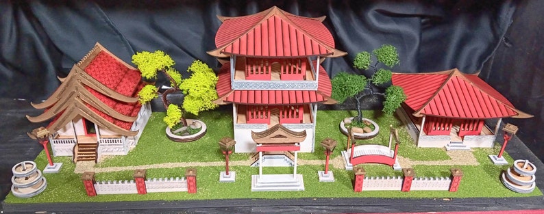 May include: A miniature model of a traditional Chinese garden with three red and white buildings, a bridge, and green trees. The buildings have intricate details and are surrounded by a green lawn.