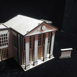 Puede incluir: Maqueta de madera de un edificio con reloj, columnas y ventanas. Un pequeño letrero junto al edificio dice "TOWN HALL". El modelo está hecho de madera clara con detalles marrón oscuro, mostrando detalles arquitectónicos.