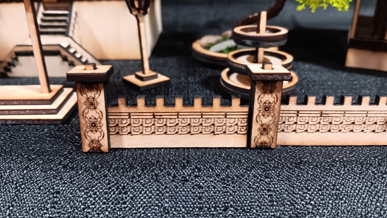 May include: A wooden miniature wall with intricate carvings and two decorative pillars. The wall is made of light brown wood and has a detailed pattern of squares and circles. The pillars are dark brown and have a more ornate design.