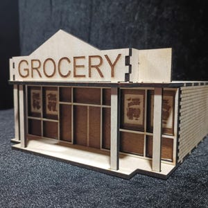 May include: A miniature wooden grocery store model with the word "GROCERY" cut out on the sign. The building features a detailed facade with windows and a brick-like side. The model is made of light-colored wood.
