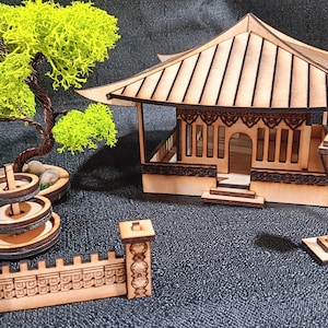 May include: A wooden model of a Japanese garden with a pagoda, a fountain, a tree, and a fence. The model is made of laser-cut wood and is painted in natural wood tones.