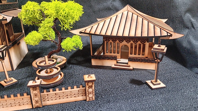 May include: A wooden model of a Japanese garden with a pagoda, a fountain, a tree, and a fence. The model is made of laser-cut wood and is painted in natural wood tones.