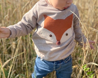 May include: A tan long-sleeved shirt featuring a large fox face applique. The fox has a textured orange knit face, cream-colored accents, and button eyes. A small, dark pink pom-pom serves as the nose. The shirt is paired with blue jeans.