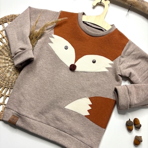 Könnte beinhalten: Ein beigefarbenes und orangefarbenes Sweatshirt mit einem Fuchsmotiv. Das Sweatshirt hat ein braunes Fuchsmotiv mit weißen Akzenten und einer kleinen Bommel-Nase. Die Ärmel sind beige und der Fuchsschwanz befindet sich unten rechts.