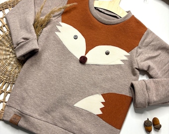 May include: A tan and orange sweatshirt with a fox face design. The sweatshirt features a brown fox face with white accents and a small pom-pom nose. The sleeves are tan, and the fox tail is on the lower right side. The sweatshirt is hanging on a wooden hanger.
