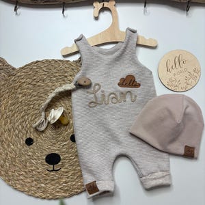 Könnte beinhalten: Ein beiger Baby-Strampler mit dem Namen "Lian" auf der Vorderseite, eine passende Mütze und ein Schnuller. Ein Holzschild mit "hello world" ist ebenfalls vorhanden. Der Strampler hängt an einem Holzkleiderbügel.
