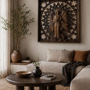 May include: A dark brown wooden angel wall art with intricate details hangs above a beige sofa. The angel is carved in a classical style, holding a staff. A round wooden coffee table and decorative vases complete the living room setting.