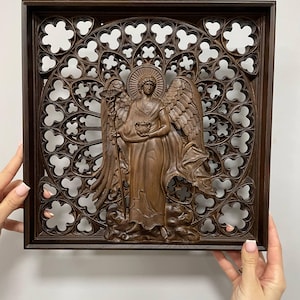 May include: A brown wooden angel sculpture with detailed wings, holding a bowl and staff, set against a decorative, openwork background within a square frame. The angel is carved in a classical style.