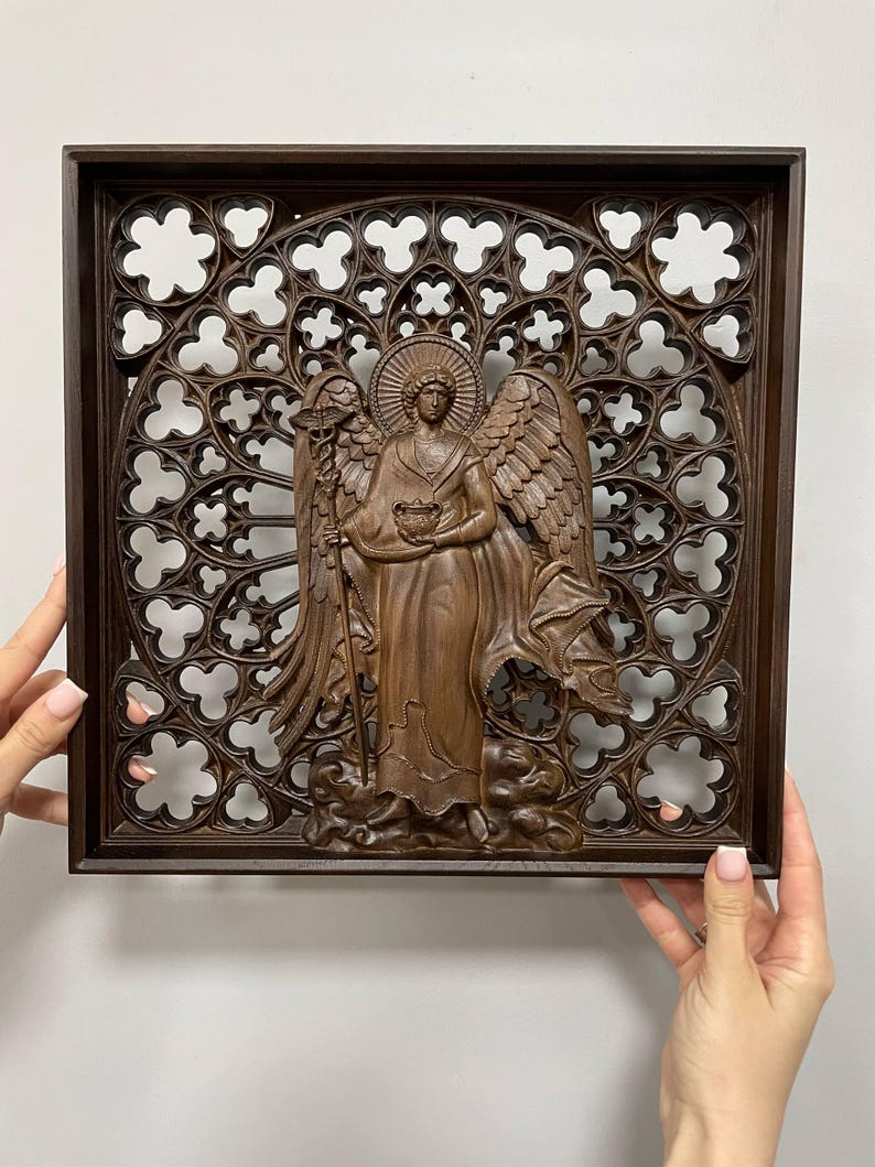 May include: A brown wooden angel sculpture with detailed wings, holding a bowl and staff, set against a decorative, openwork background within a square frame. The angel is carved in a classical style.