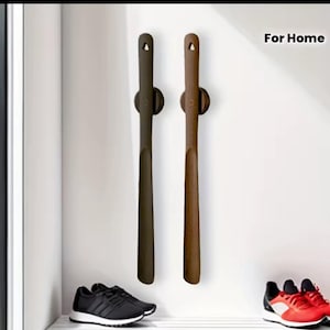 May include: Two shoe horns, one dark brown and one light brown, are mounted on a white wall. The shoe horns are long and slender, with a small hole at the top for hanging. The text "For Home" is in the upper right corner.
