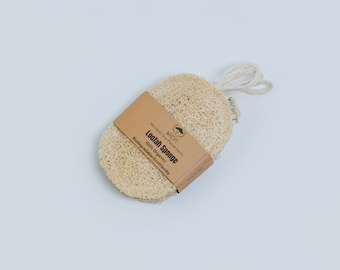 May include: A natural loofah sponge with a woven texture, featuring a brown paper band with the text "Lootah Sponge 100% Organic". The sponge has a loop for hanging and is presented on a white background. Eco-friendly and biodegradable.