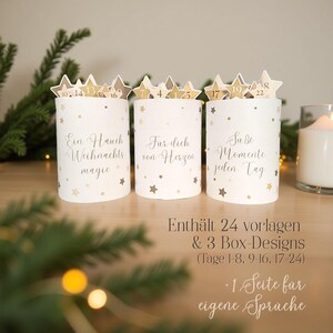 Op de afbeelding: Drie witte cilindrische adventskalenderdozen met gouden steraccenten en stervormige toppers met nummers. Elke doos heeft een andere inscriptie in cursief. De tekst op de dozen luidt: "Ein Hauch Weihnachts magie", "Fur dich von Herzen" en "Sube Momente jeden Tag".