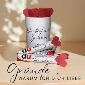 Könnte beinhalten: Ein weißer zylindrischer Behälter gefüllt mit roten Papierherzen, mit dem Text "Du bist mein Zuhause". Darunter drei verpackte Süßigkeiten mit roten Herzakzenten und Text. Die Worte "Gründe, warum ich dich liebe" stehen unten.