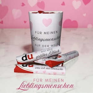 Pode incluir: Uma caneca branca com corações rosa e texto em alemão, incluindo "FÜR MEINEN Lieblingsmenschen". Várias barras de chocolate com texto em alemão, incluindo "du bist mein größtes Glück".