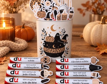 May include: A cylindrical container decorated with a black cauldron, ghost, and moon illustration, filled with assorted die-cut paper shapes. Eight white candy bars are arranged in front, each featuring the text du and German phrases.