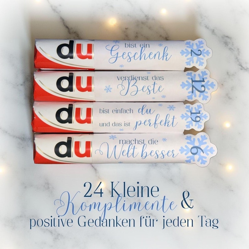 May include: Four chocolate bars with white wrappers and red accents. Each wrapper has a different message in blue script, with snowflake accents. The text "24 Kleine Komplimente & positive Gedanken f&uuml;r jeden Tag" is also visible.