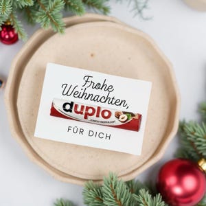 May include: A white card with the text "Frohe Weihnachten, Duplo FÜR DICH" and a Duplo chocolate bar. The card is on a beige plate, surrounded by Christmas decorations including pine branches and a red bauble.