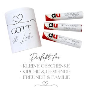 May include: A white cylindrical gift box with the words "GOTT ist Liebe" in black script, accompanied by three candy bars. The candy bars have red and white packaging with German text. The image also includes text that says "Perfekt f&uuml;r" and lists gift ideas.