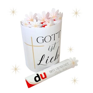 May include: A white cylindrical container with the words "GOTT ist Lieb" and a gold cross. The container is filled with white flower-shaped decorations. A white candy bar with the word "du" and the text "BIST GESEGNET" is next to the container.