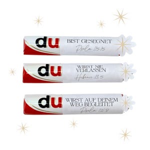 May include: Three chocolate bars with white wrappers and red accents. Each wrapper has the word "du" in black, and a different German phrase with a Bible verse reference. Decorative starbursts are on the right.