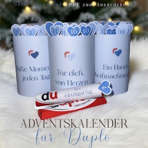 May include: Advent calendar for Duplo candies. The calendar features three light blue cylindrical containers with heart-shaped cutouts numbered for each day. The containers have text in German. A Duplo candy bar is placed in front.