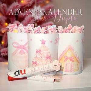 May include: A white advent calendar with pink illustrations of a Christmas tree, gingerbread house, and ornaments. The calendar has numbered compartments and includes a chocolate bar and a small paper with text.
