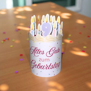Pode incluir: Decoração branca para bolo de aniversário com o texto "Alles Gute zum Geburtstag" em escrita bordô. O topo do bolo apresenta o número 9 e várias velas com chamas amarelas. Confetes coloridos espalhados.