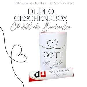 May include: A white gift box with a heart design and the words "GOTT ist Liebe" printed on it. A wrapped chocolate bar with the word "du" and the text "BIST GESEGNET Psalm 115:15" is in front of the box. The image also includes the text "DUPLO GESCHENKBOX Christliche Banderolen".