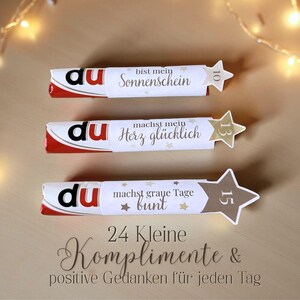 Op de afbeelding: Drie chocoladerepen met witte verpakkingen en rode accenten. Elke verpakking heeft een Duitse zin en een stervormig label met een nummer. De tekst bevat "Sonnenschein", "Herz gl&uuml;cklich" en "graue Tage bunt".