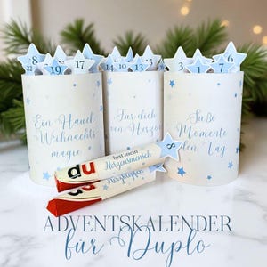 Può includere: Calendario dell'Avvento per cioccolatini Duplo. Contenitori cilindrici bianchi con dettagli a stella blu e etichette numeriche a forma di stella. I contenitori hanno testo in tedesco e le confezioni di cioccolato sono visibili.