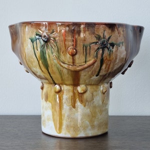 Peut inclure: Un grand bol en céramique avec une base large et arrondie et un bol large et peu profond. Le bol est décoré d'un glaçage brun, jaune et vert, et présente un motif de visage souriant stylisé.