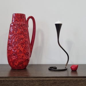 May include: A red ceramic vase with a textured surface and a black metal candle holder with a white candle on a brown wooden surface.
