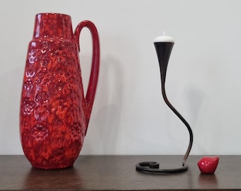 May include: A red ceramic vase with a textured surface and a black metal candle holder with a white candle on a brown wooden surface.