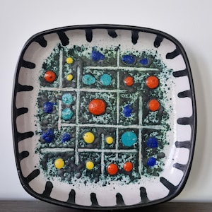 Può includere: Piatto quadrato in ceramica con bordo nero e bordo bianco con linee nere verticali e orizzontali. Il centro ha uno sfondo verde maculato con punti colorati arancioni, blu, gialli e turchesi.